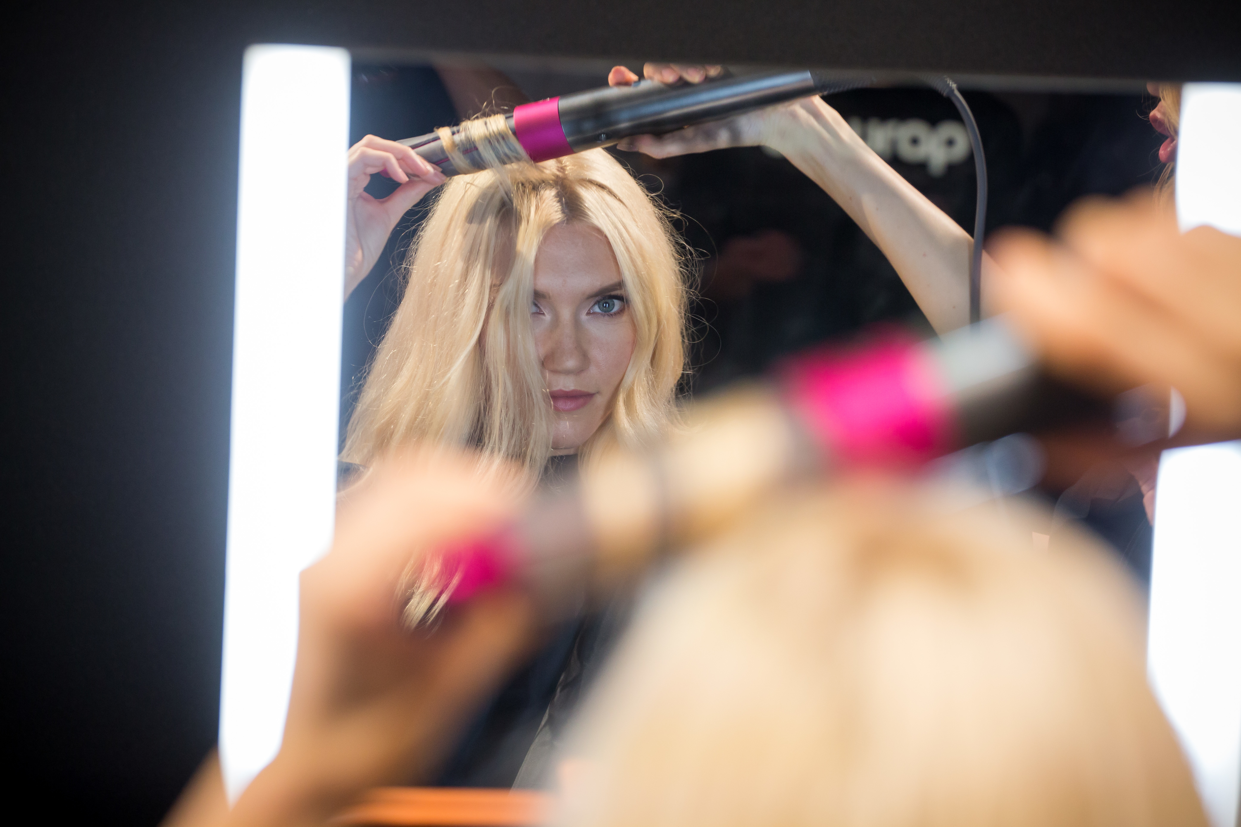 A woman styles her hair in front of a mirror using a curling tool. Her reflection is visible as she curls a section of her long hair