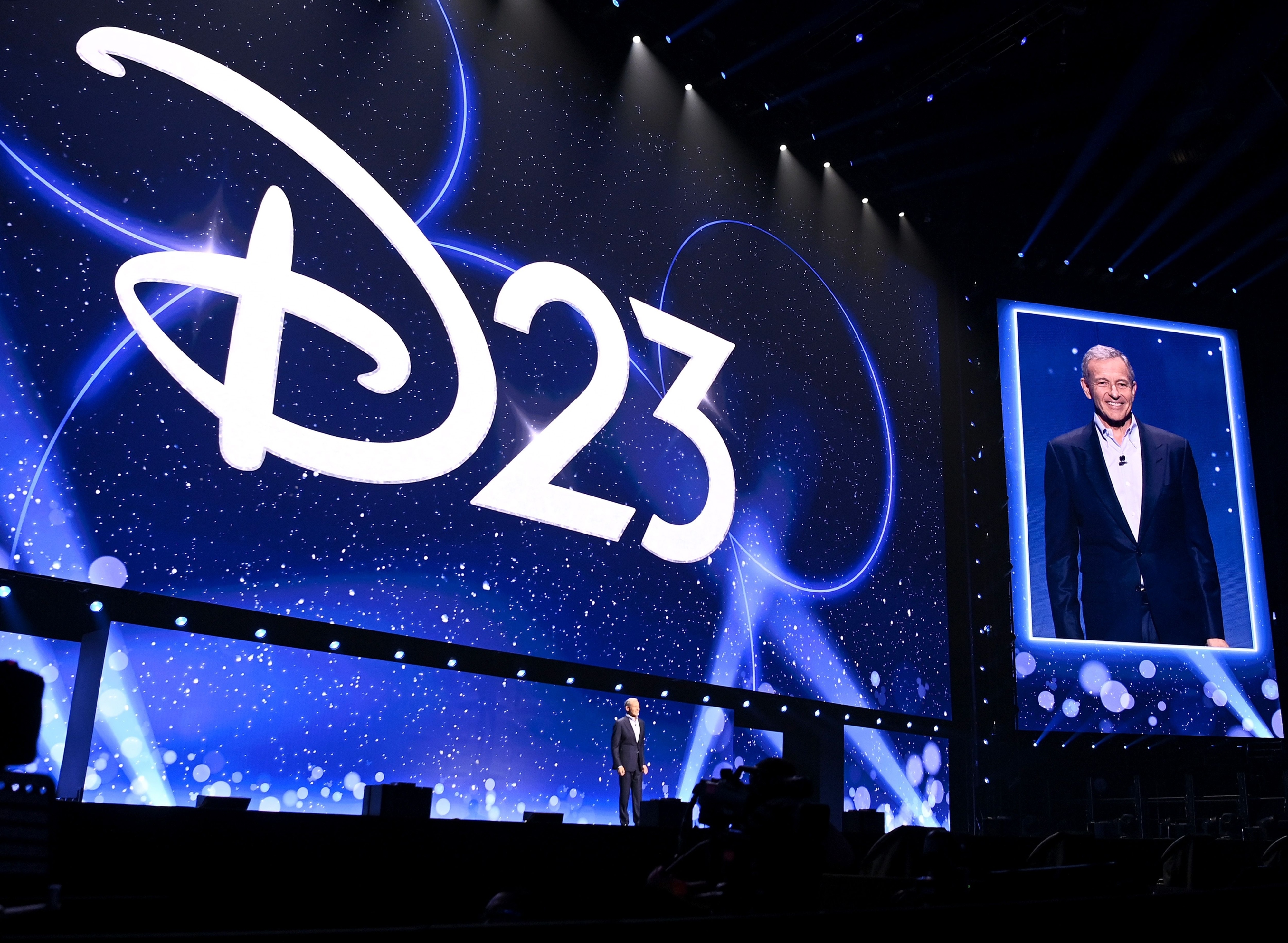 Man speaking on stage at D23 Expo, large D23 logo on screen behind him