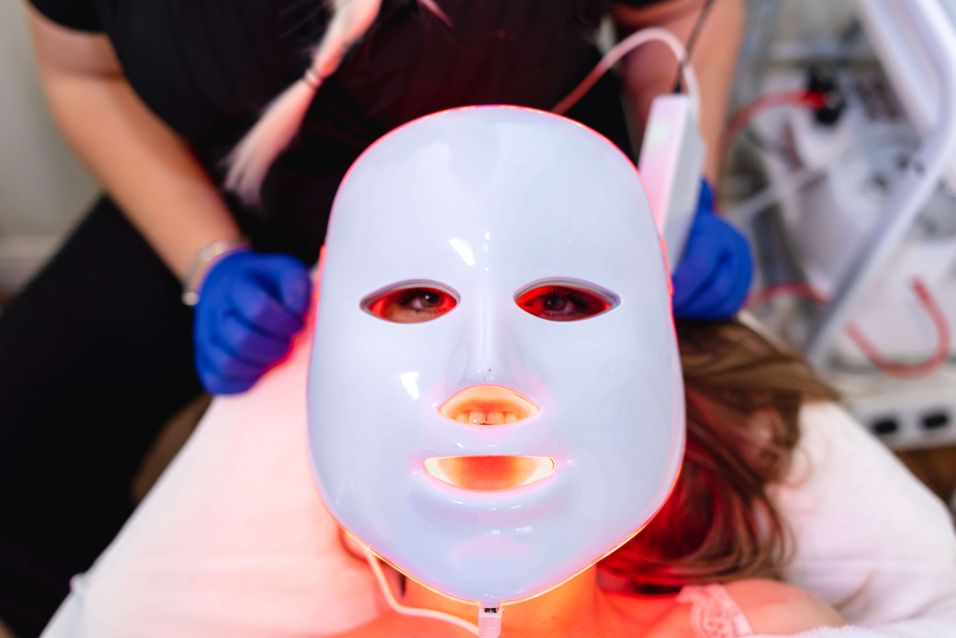 Person lying down wearing a glowing LED facial mask, attended by someone with blue gloves