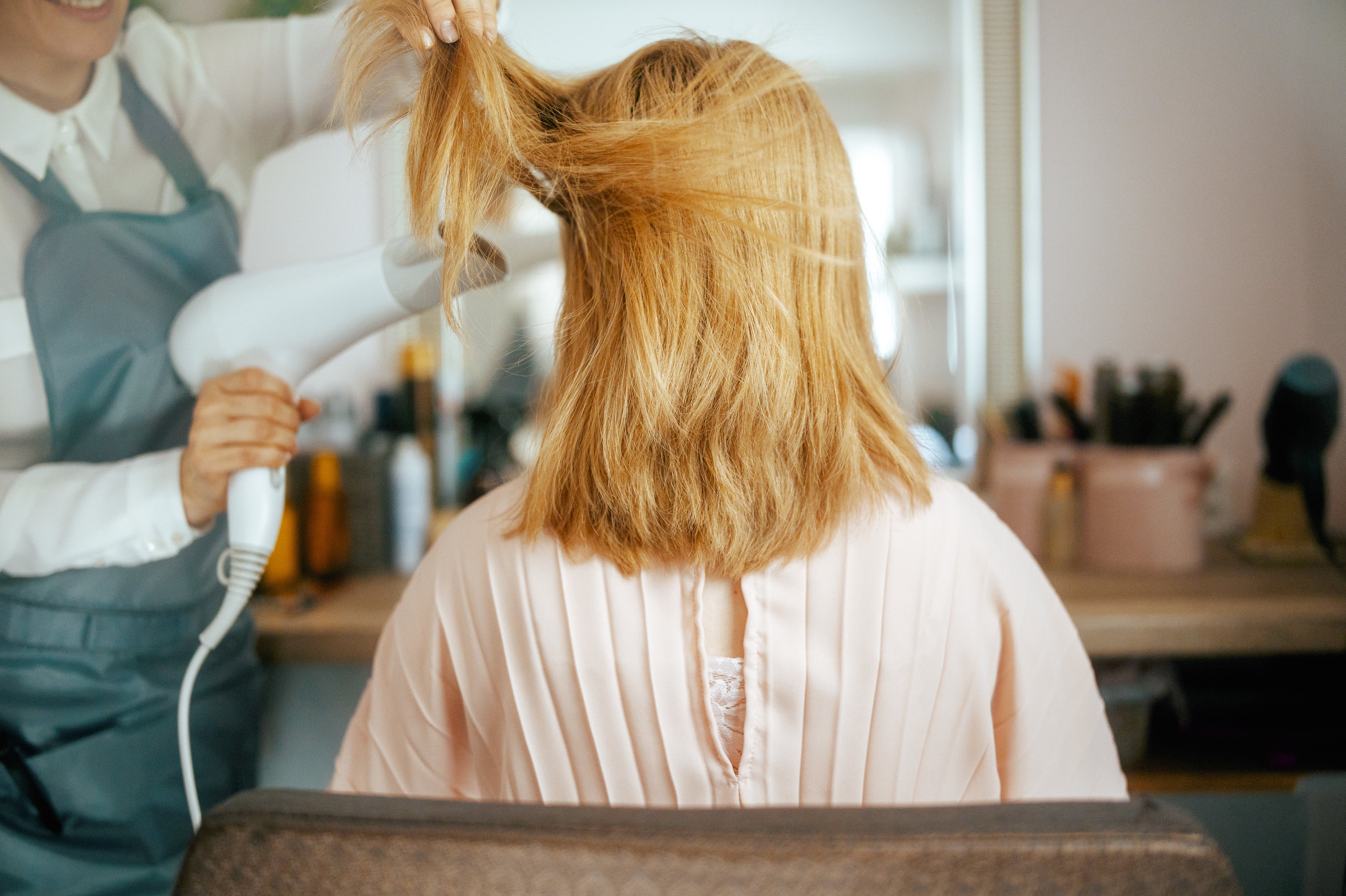 Person with medium-length hair getting a blow-dry in a salon. Stylist uses a hair dryer