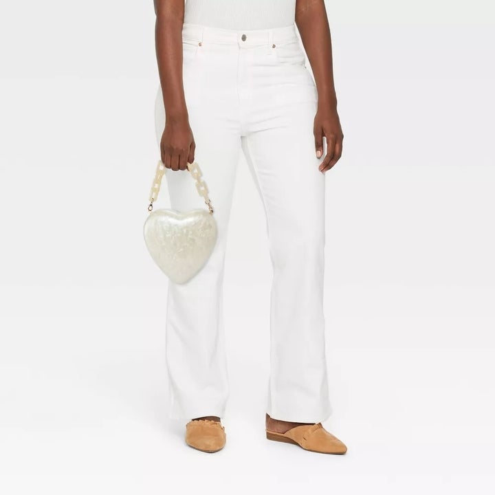 Person wearing white flare jeans, holding a pearlescent heart-shaped handbag, and wearing tan loafers