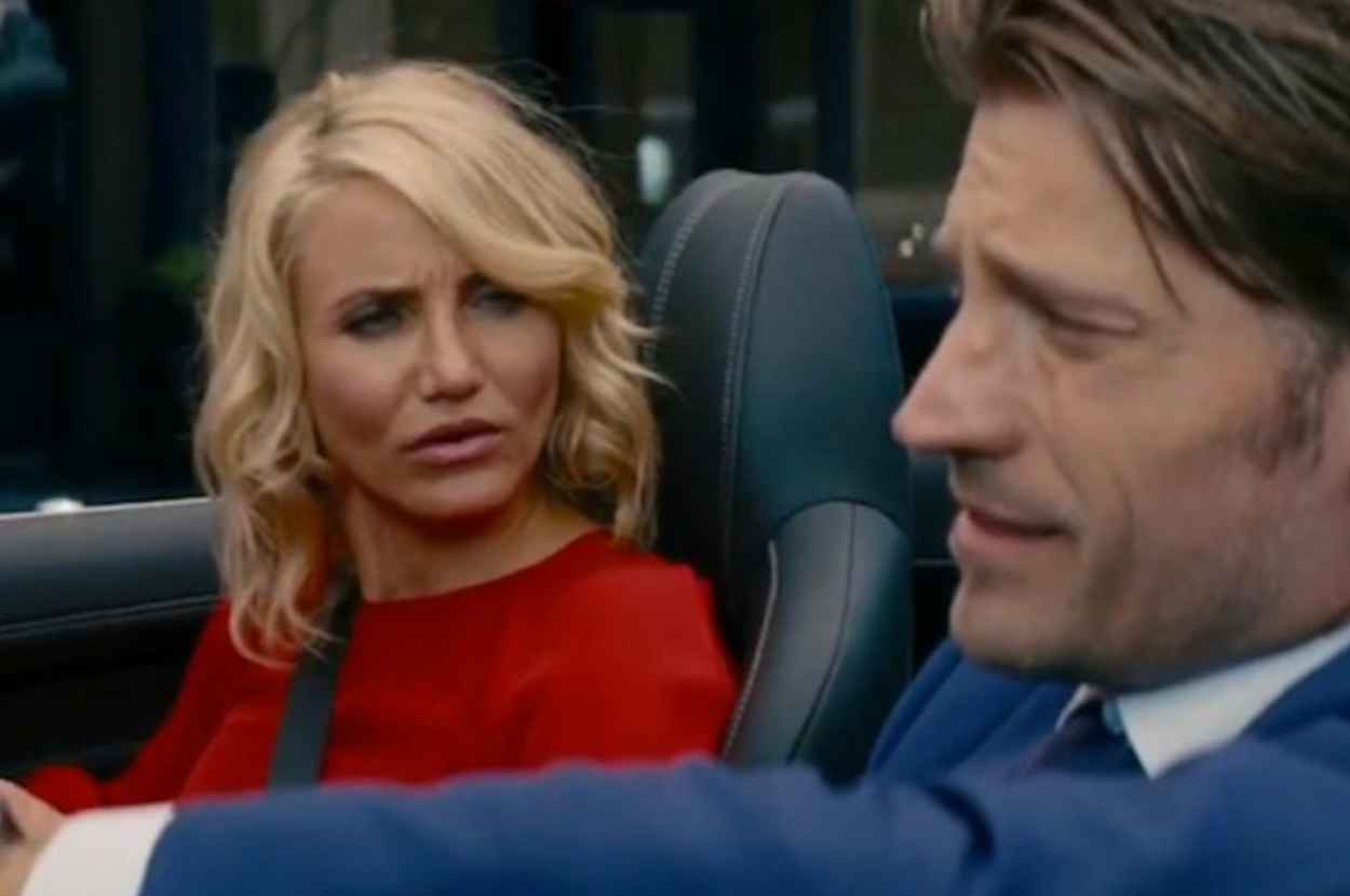 Two people sit in a car. The woman has wavy hair and wears a stylish red outfit, while the man wears a formal suit, both engaged in conversation
