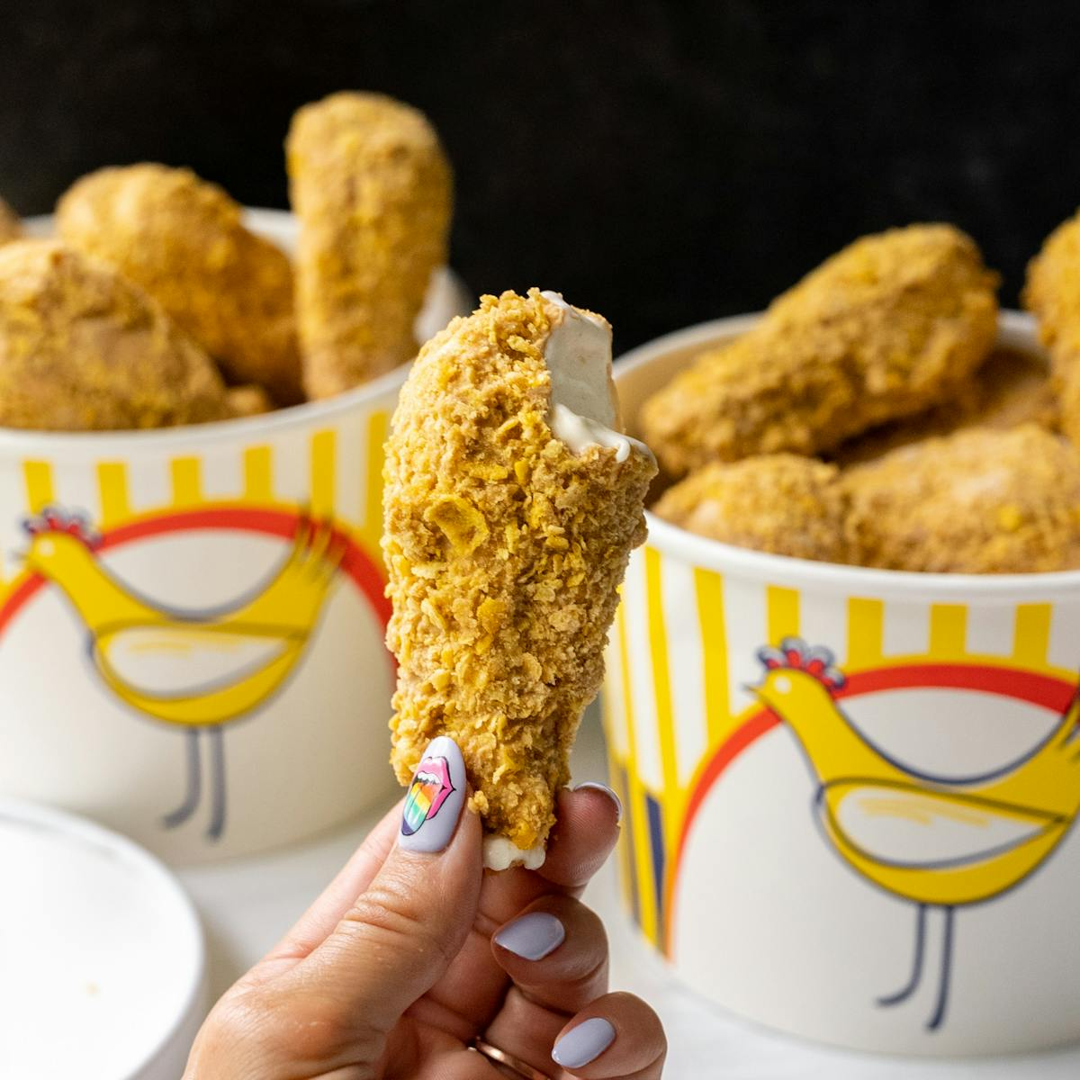 Hand holding a drumstick-shaped ice cream cone covered in crushed cornflakes, with chicken buckets in the background