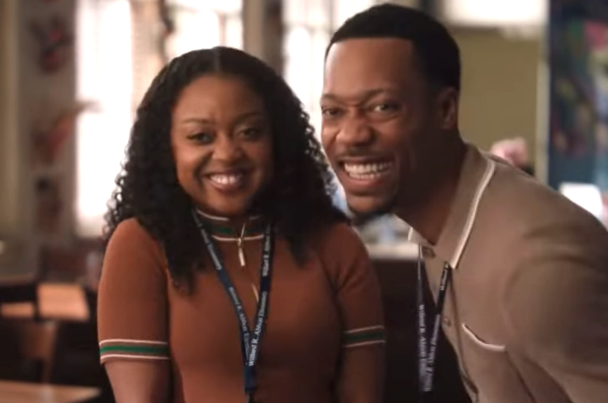 Two people smiling, wearing lanyards; one in a brown shirt, the other in a beige top. They're in an indoor setting, appearing happy together