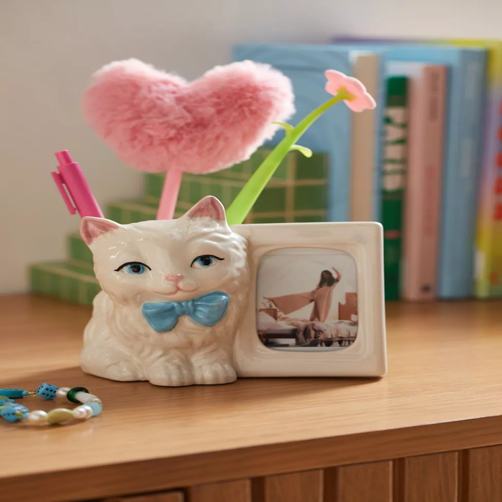 Cat pen holder with photo frame, heart pen, and books in the background on a wooden desk