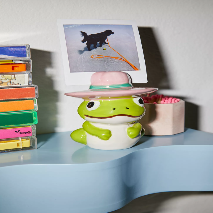 Ceramic frog with a pink hat holds a photo of a dog