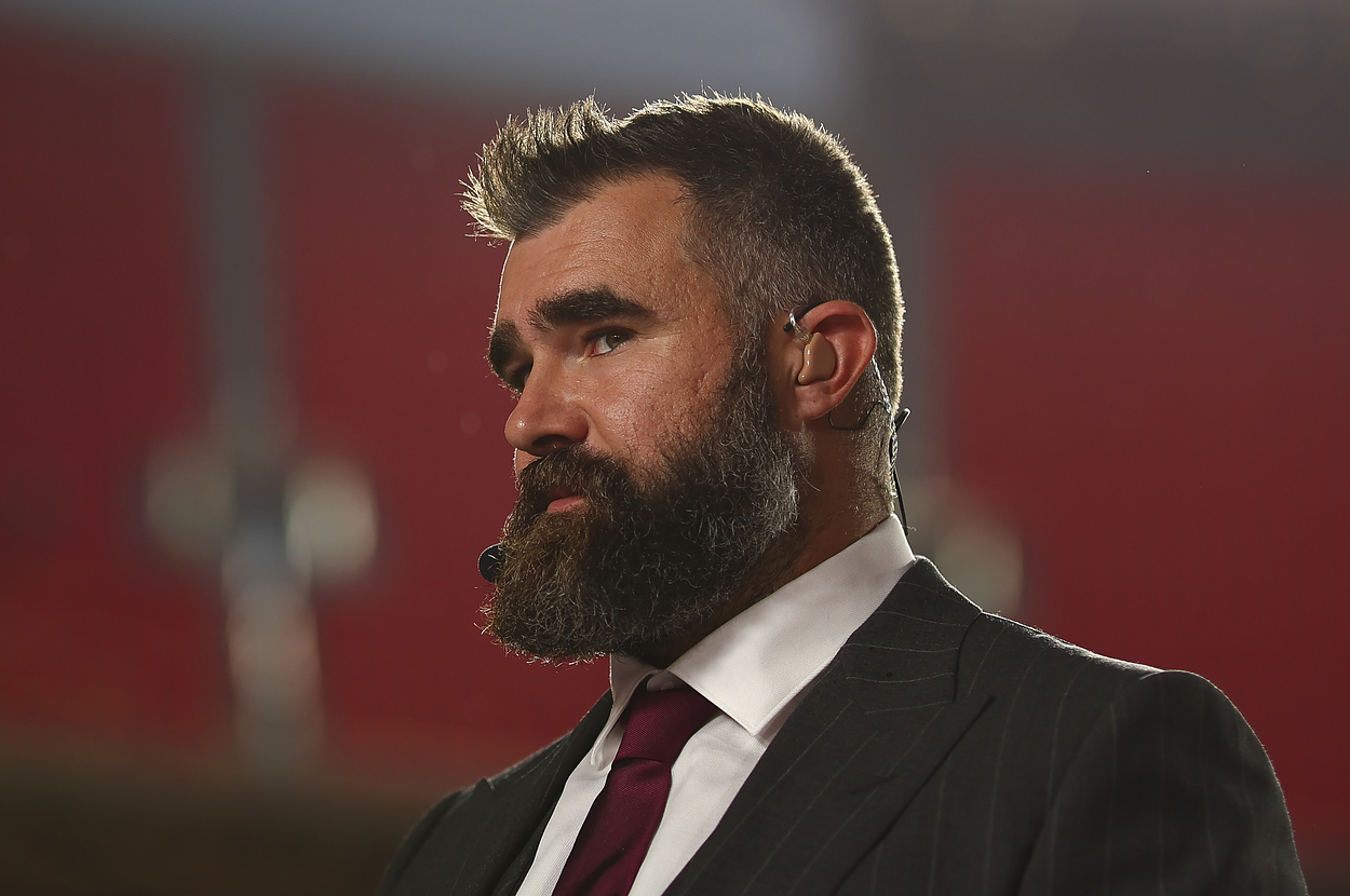 A man with a beard and wearing a suit and tie stands on a red carpet, wearing a headset microphone, in a well-lit venue