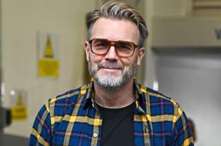 Gary Barlow wearing sunglasses, a plaid shirt over a black t-shirt and jeans, standing in a casual pose with hands in pockets