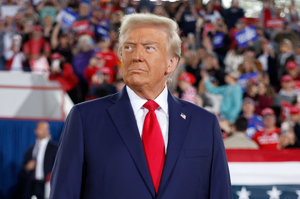A man in a suit stands at a political rally with a large crowd and American flag-themed decorations in the background