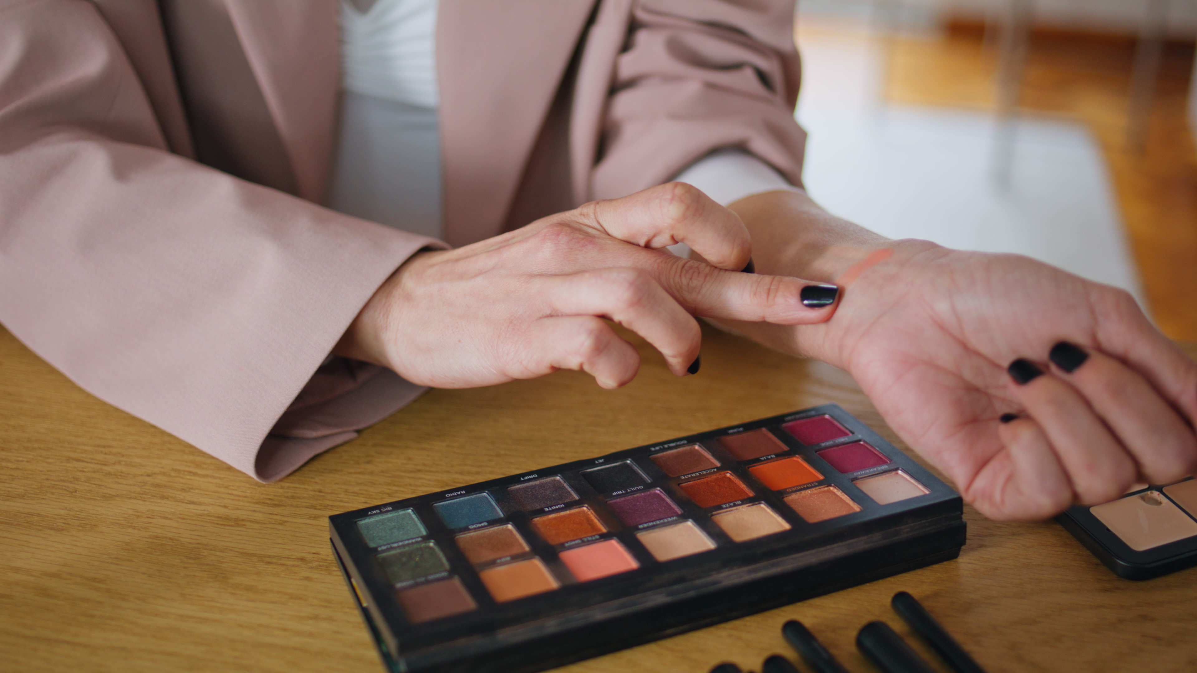 Person testing makeup shades on wrist from an eyeshadow palette on a wooden table