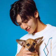 A person holding a small dog, smiling gently. The person is wearing a white sweater with a simple stitched pattern