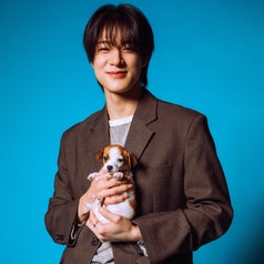 Person in a blazer and jeans smiles while holding a small puppy in front of a blue background