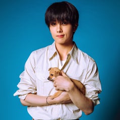 Person holding a small puppy, wearing a white shirt and tan pants, against a blue background