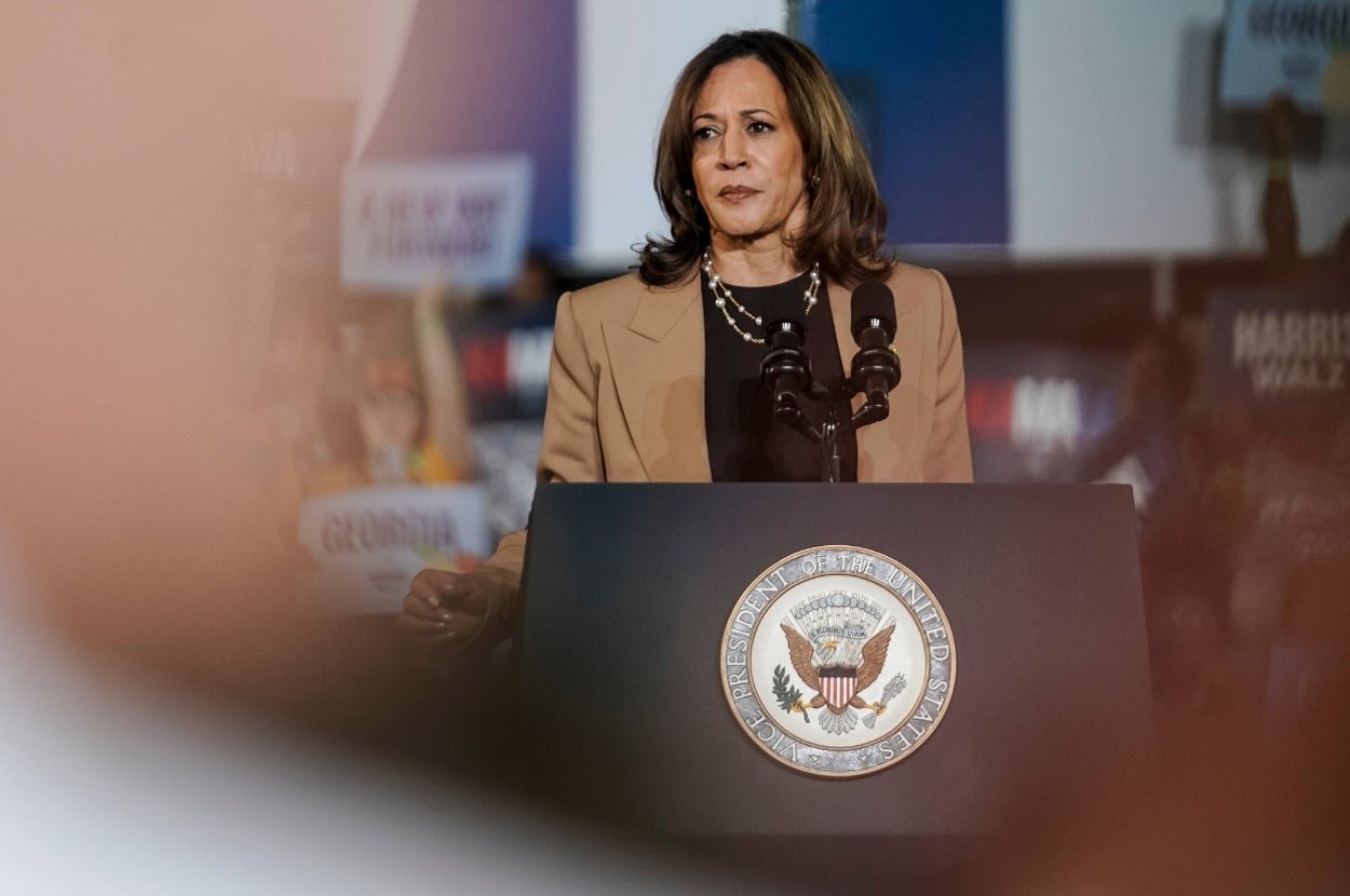 A woman stands at a podium with the presidential seal, addressing an audience. She's wearing a tan blazer over a black top