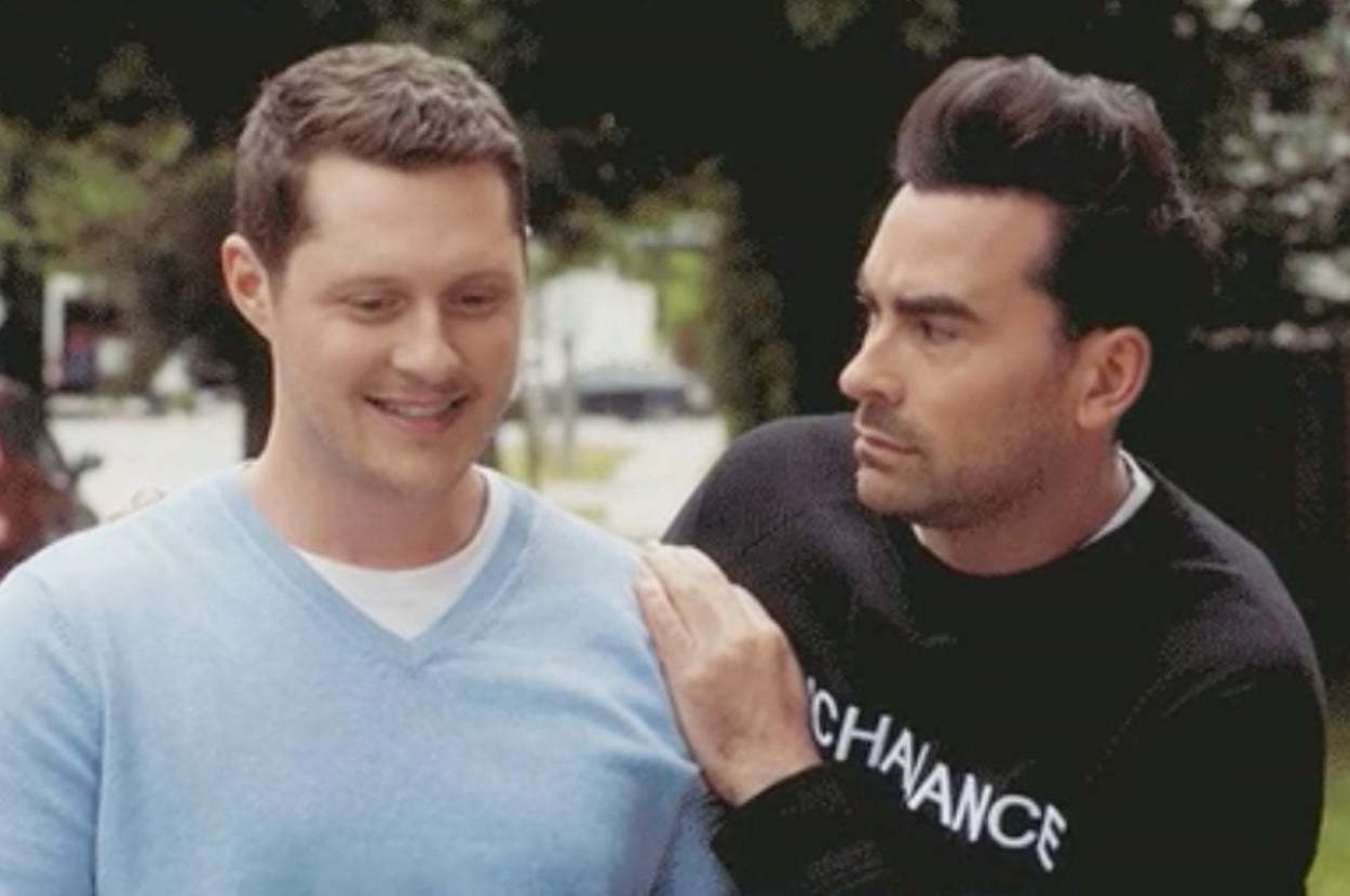 Two men sharing a light moment outdoors. One smiles while the other, with styled hair and a sweatshirt, playfully rests a hand on his shoulder