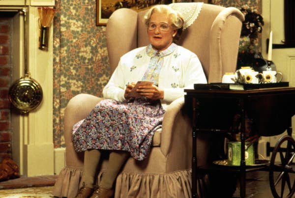 Robin dressed as an elderly woman, wearing a floral dress and cardigan, sits in a vintage armchair, holding a cup