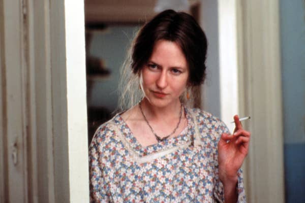 Nicole wearing a floral-patterned dress, holding a cigarette, standing in a doorway with a pensive expression