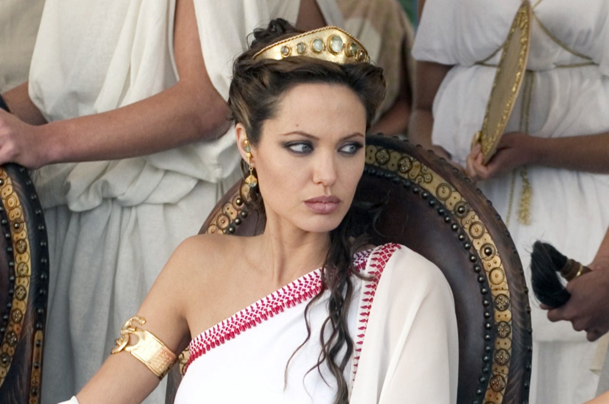Angelina Jollie in ancient-style white toga and crown, seated on a chair, looking to the side. Others in similar attire are in the background