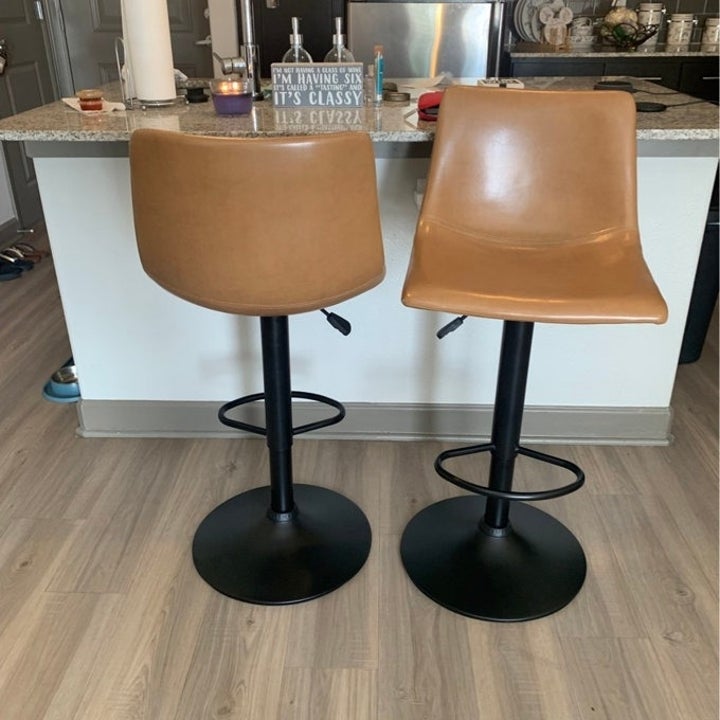 Two modern-style barstools with tan seats and black bases in a kitchen setting. Countertop and decor items are visible in the background