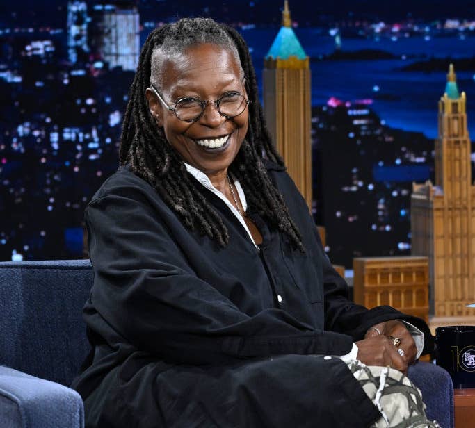 Person with dreadlocks, smiling, seated on a talk show set, wearing a black shirt and patterned pants. City skyline backdrop visible