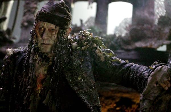 Stellan in tattered clothing with seaweed-like attire, resembling a weathered pirate, against a dark, ship-like backdrop from a movie scene