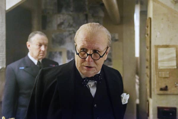 Gary portraying Winston Churchill wears a suit with a bow tie and round glasses, standing in a room with a serious expression