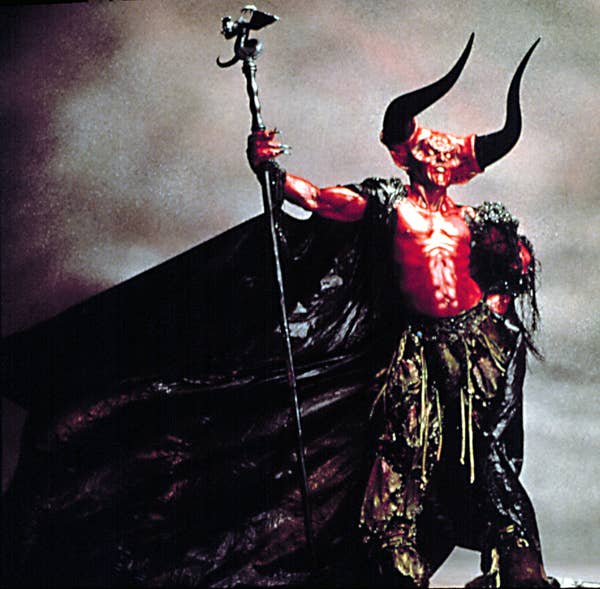 Tim in an elaborate fantasy costume with large horns and a cape, holding a staff, stands dramatically against a cloudy backdrop