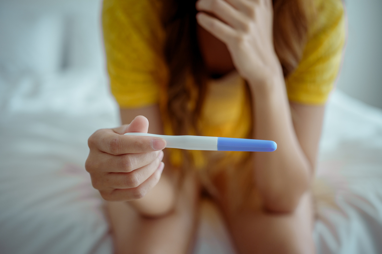 Person holding a pregnancy test while sitting on a bed, wearing casual clothing