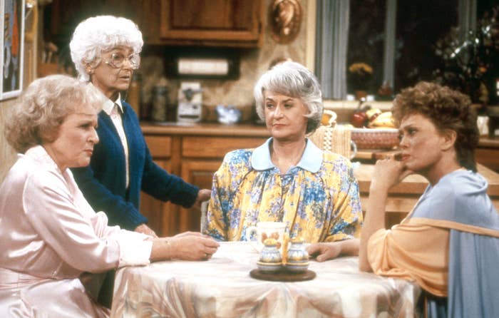 Four elderly women sit around a kitchen table, engaged in conversation. They wear vintage, elegant loungewear, reflecting a cozy domestic scene