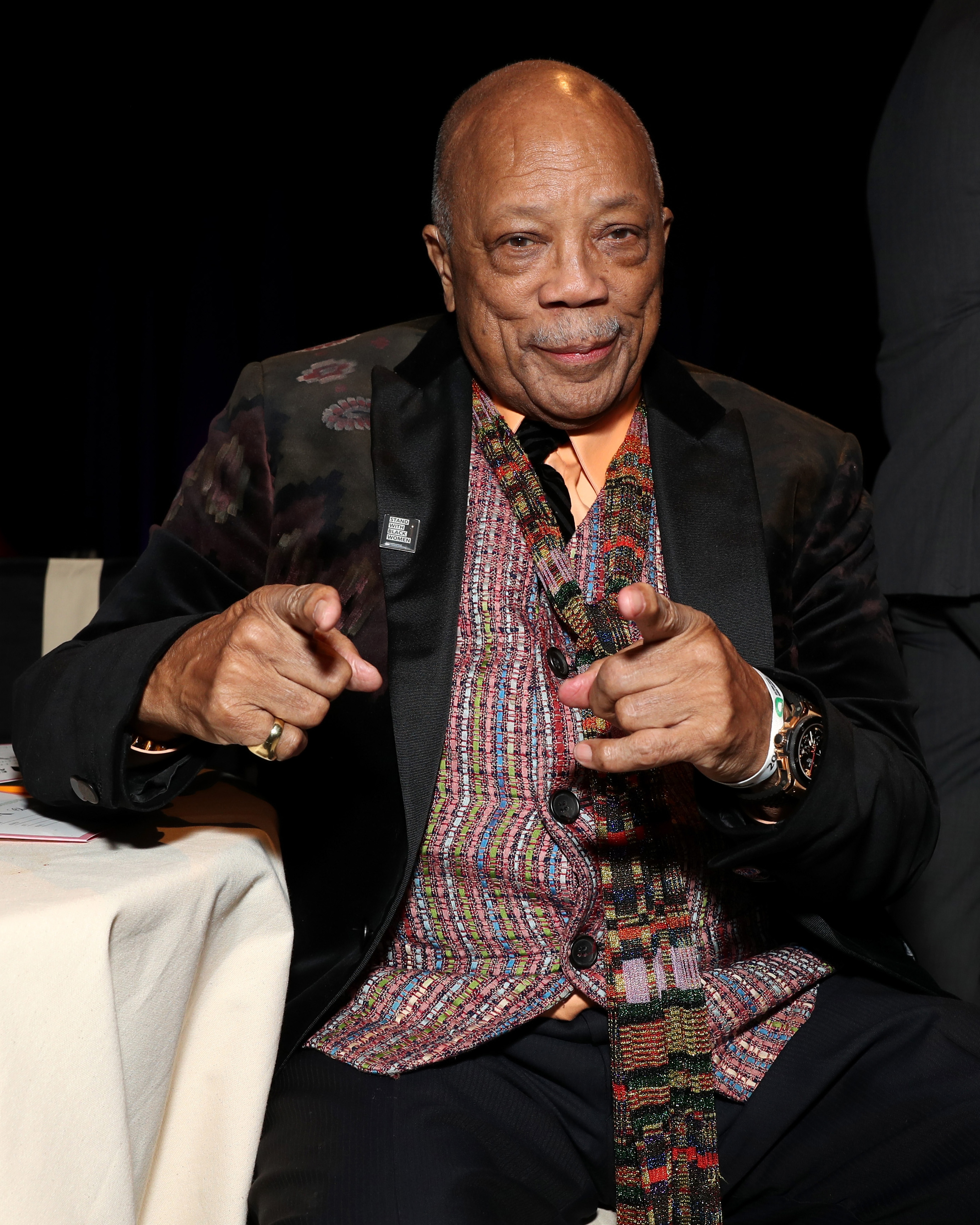 Quincy Jones n a patterned vest and jacket poses playfully with finger guns while seated at a table