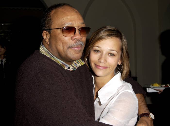 Quincy Jones wearing sunglasses and a dark sweater, Rashida Jones in a white blouse, both smiling at an event with Rashida's arms wrapped around Quincy