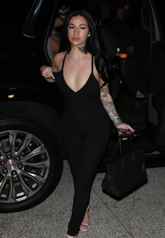 Bhad Bhabie with long hair, wearing a form-fitting outfit, holding a bag, exiting a vehicle