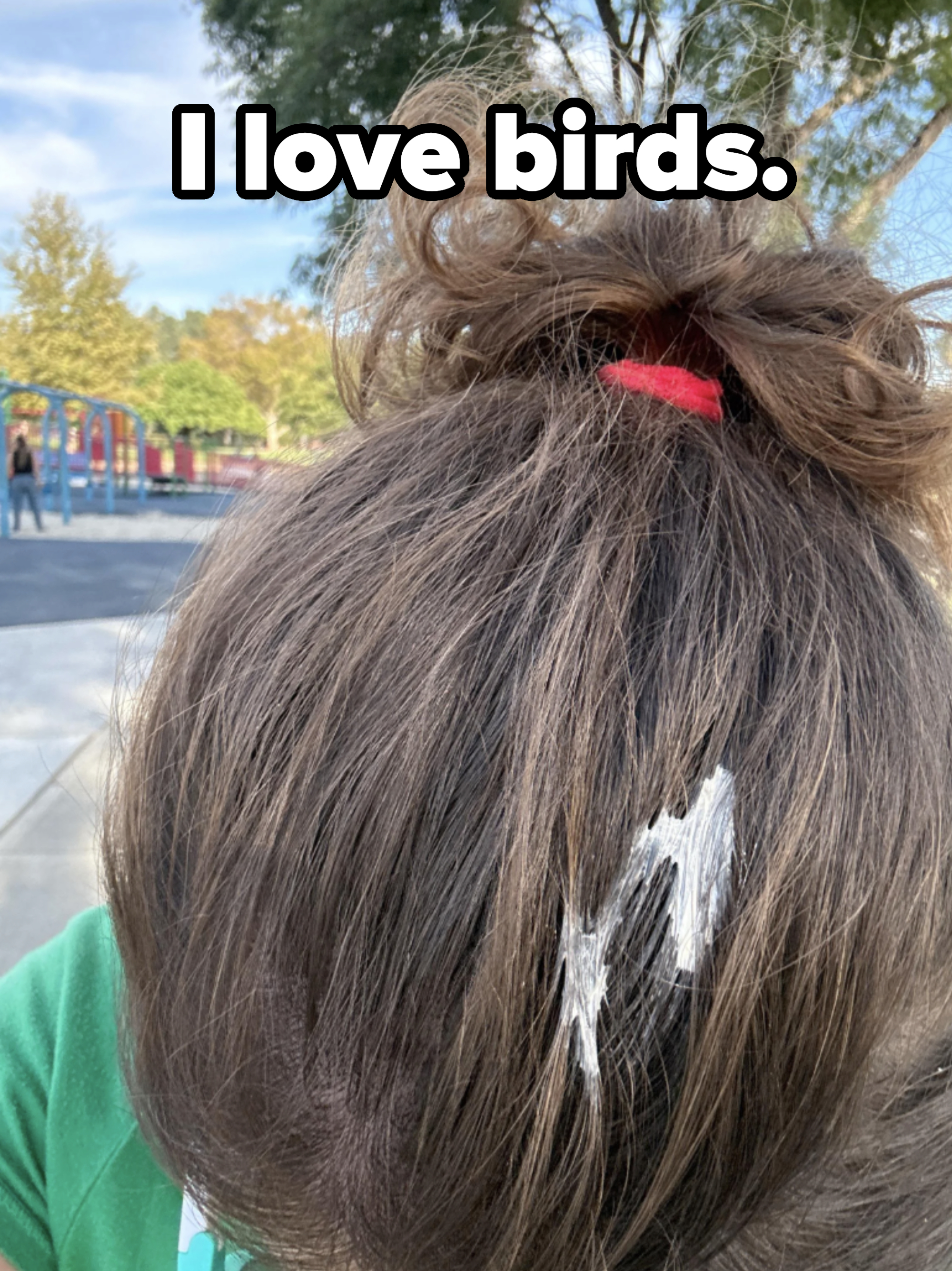 A person with hair in a bun and white paint streaks bows their head outdoors in a park setting