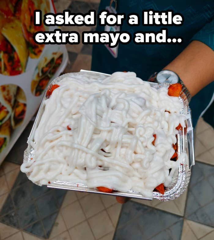 A person holds a foil container filled with fries or a similar snack covered in a generous layer of white sauce, possibly mayonnaise