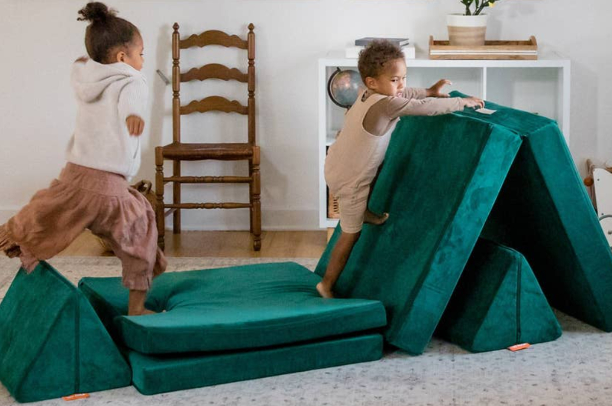 Two children play on a set of modular green foam cushions in a living room, climbing and jumping creatively