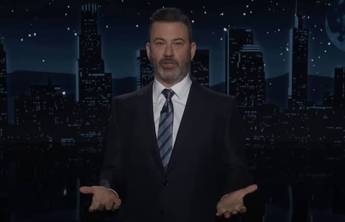 A person in a suit with a striped tie gestures with open hands against a city skyline backdrop at night