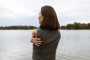 Person with shoulder-length brown hair stands by a lake, hugging themselves; viewed from the back