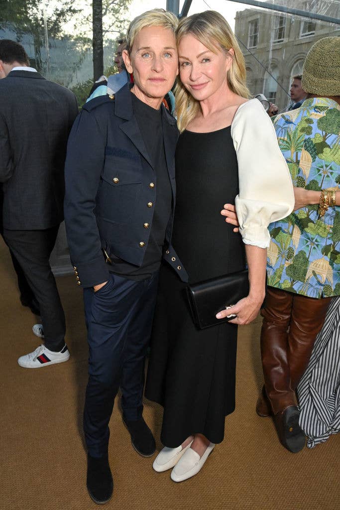 Two people posing at an event, one in a dark blazer and pants, the other in a dress with puff sleeves, both smiling and standing closely together