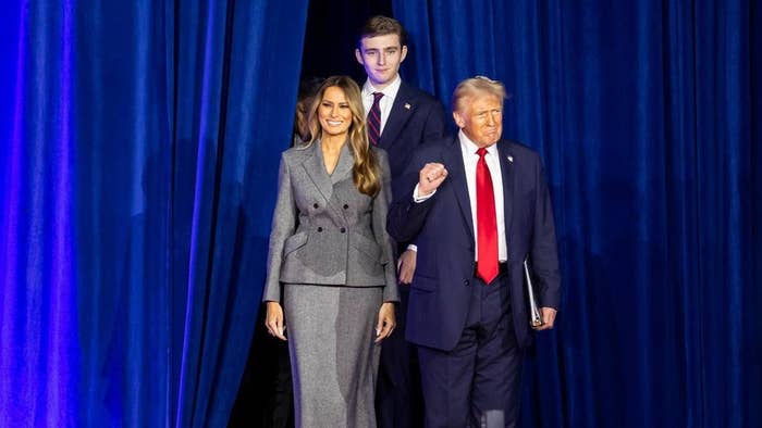 Three people walk on stage; the woman wears a tailored suit, and one man in a suit raises his fist. The context suggests a formal or political event