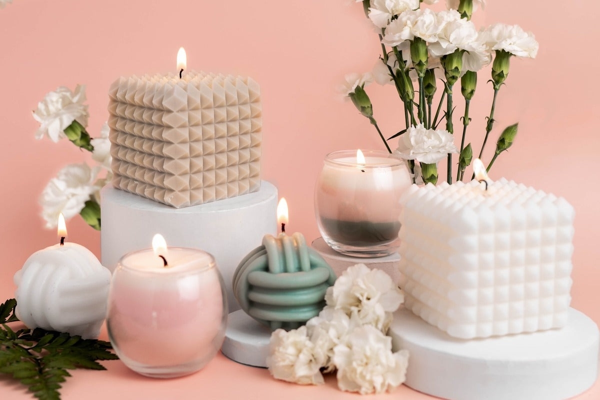 A collection of uniquely shaped and textured candles displayed with flowers on a pink background