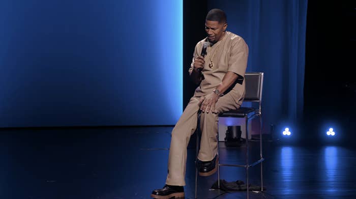Person on stage sitting on a stool, holding a microphone, wearing a casual outfit