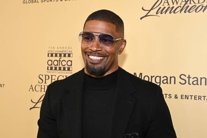 A smiling man in a black turtleneck and blazer, wearing sunglasses, attends an awards luncheon event with backdrop text