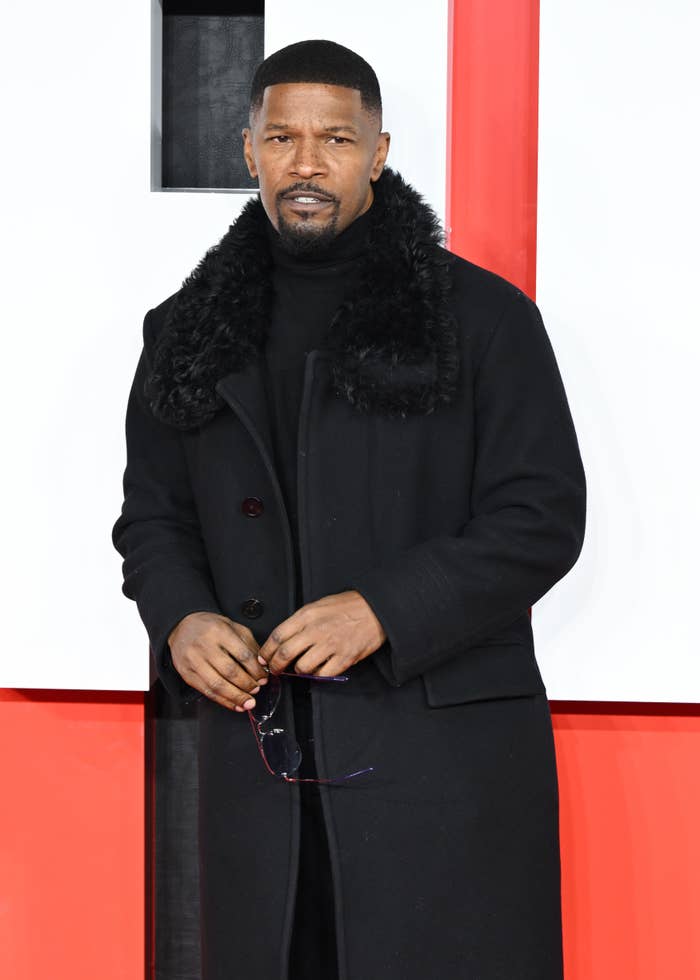 Person in a long black coat with a fur collar poses on a red carpet. They hold sunglasses and smile slightly