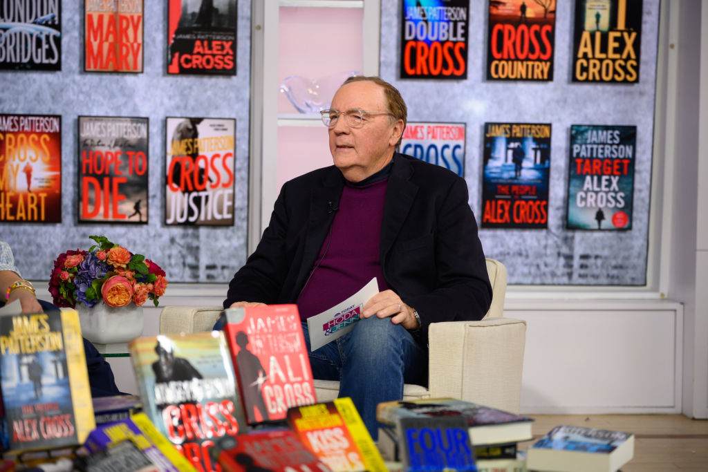 Author sitting among displayed books, holding one book, in a TV studio setting. Shelves show various book covers with the author's name