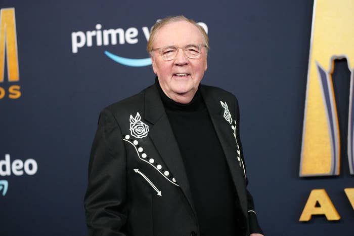 Person in a black jacket with white floral and line details, smiling at a media event backdrop