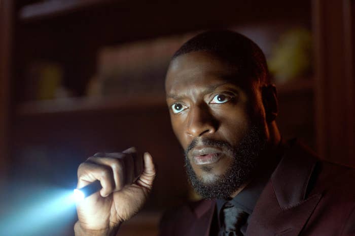 A person in a dimly lit room holds a flashlight, looking intently forward, wearing a formal suit with a serious expression