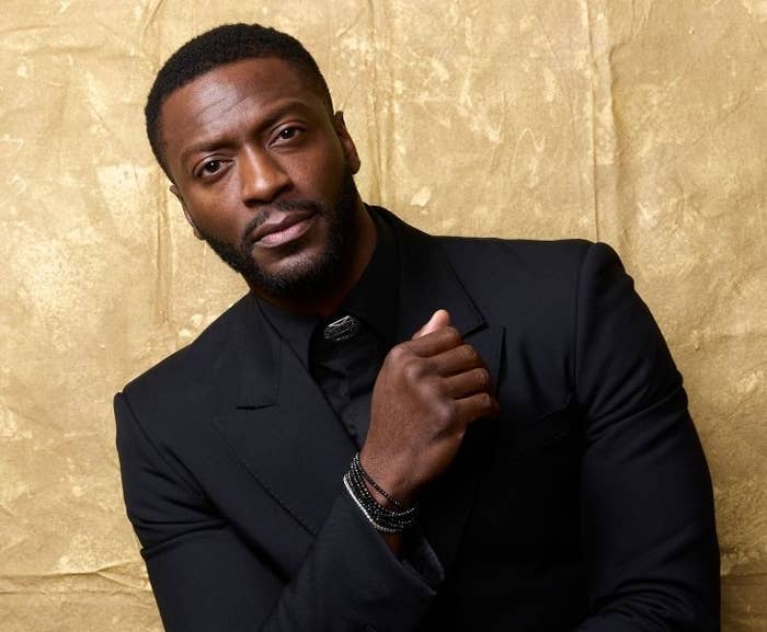 Man in a sleek black suit poses against a textured backdrop, exuding sophistication and style