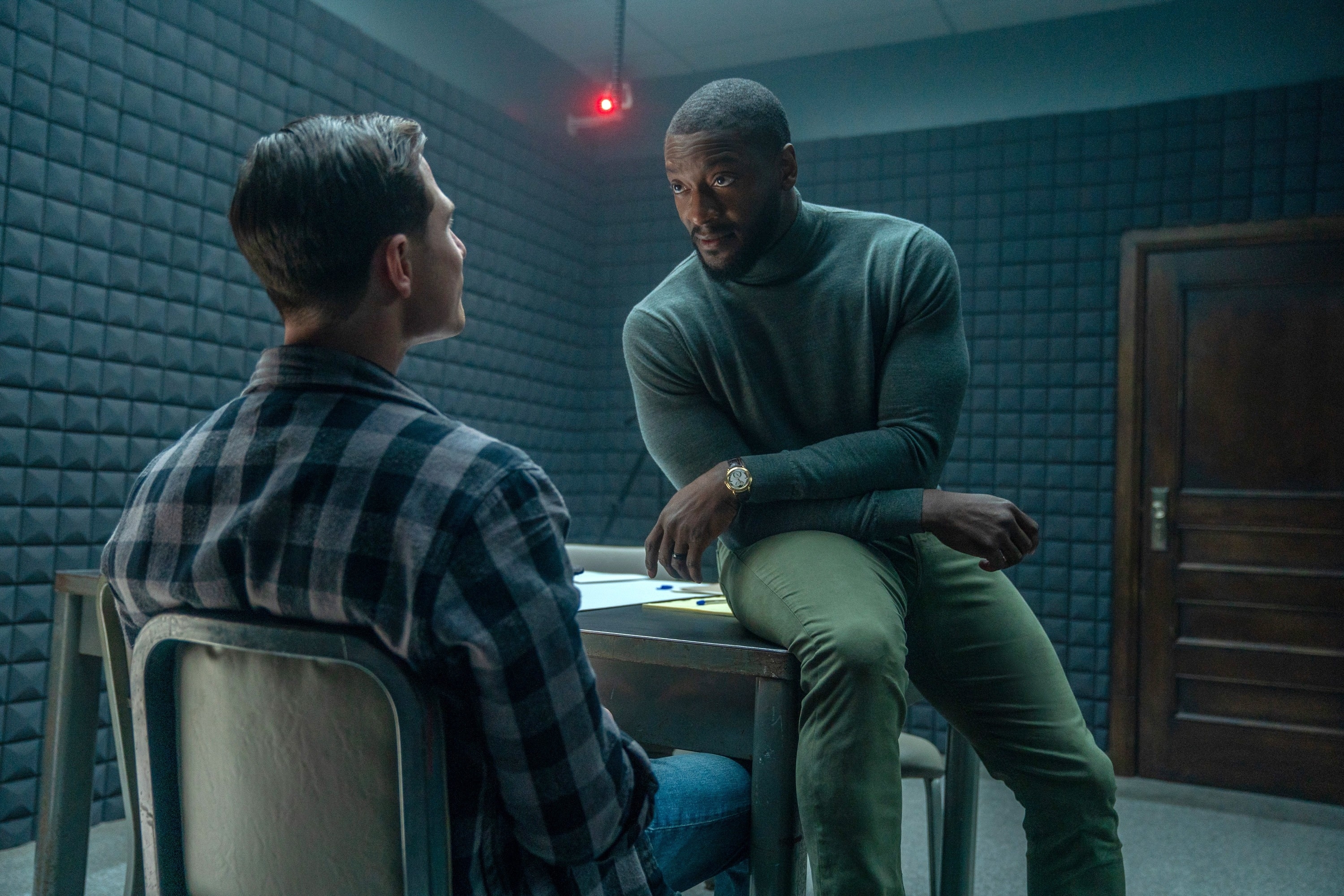 Two men in a soundproof room, one seated wearing a plaid shirt, the other perched on a table in a turtleneck and watch, engaging in a serious discussion