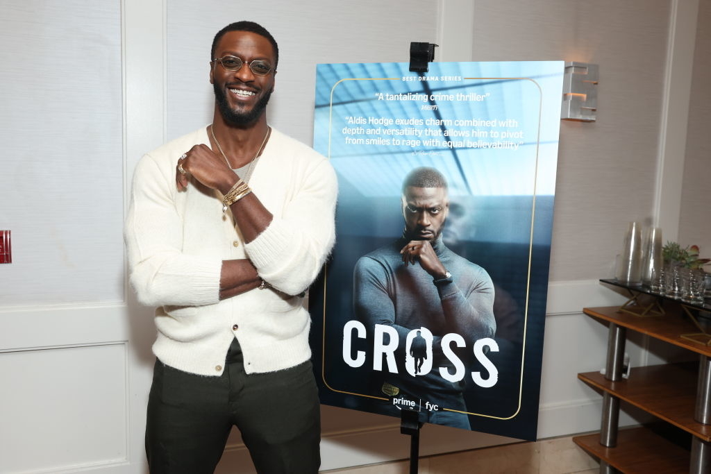 Man in a casual outfit smiling next to a movie poster that reads "Cross." The poster features the same man in a thoughtful pose