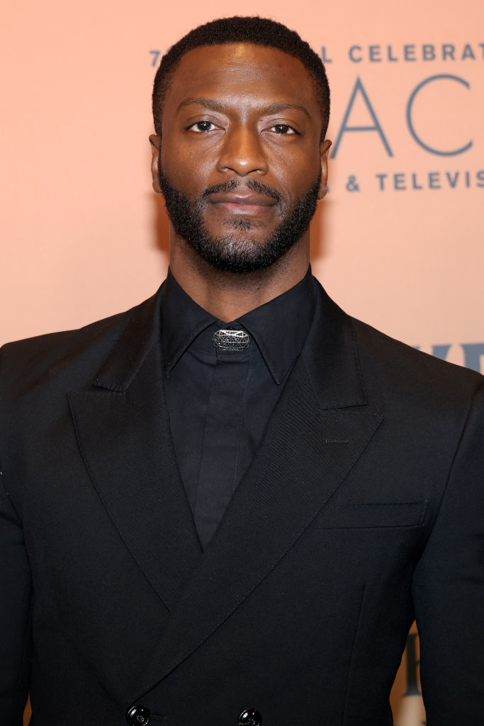 A person in a sleek black suit at a celebrity event, posing for the camera with a neutral expression